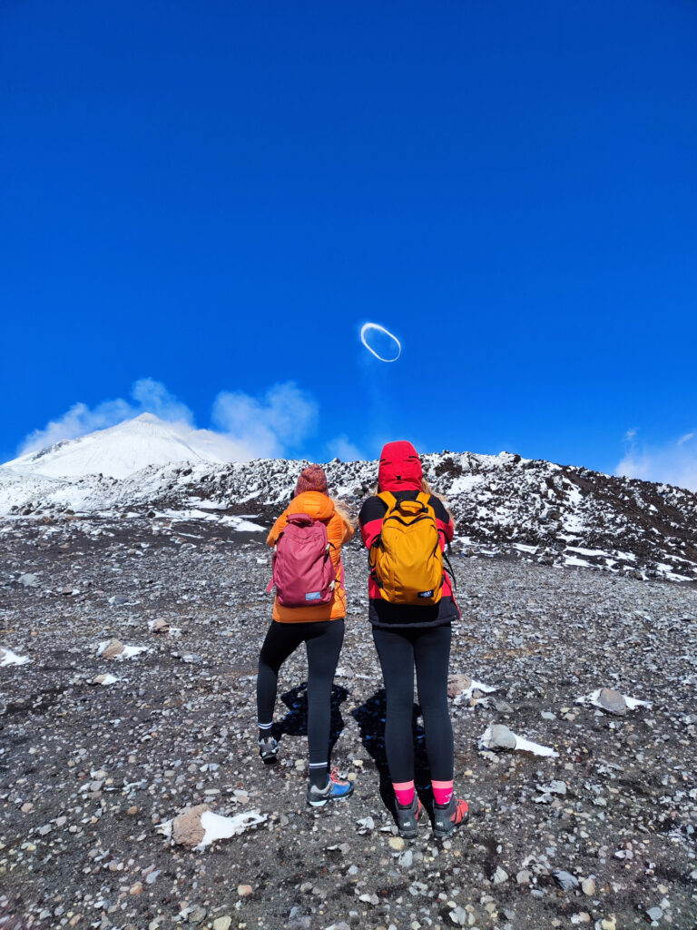 Two hikers, dwarfed by the grandeur of Mount Etna, look up at a striking vortex ring formed from volcanic emissions, a blend of science and spectacle.